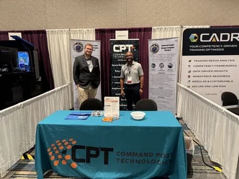 Two people standing behind a teal Command Post Technologies table at a conference booth, with CPT and CADRE banners behind them.