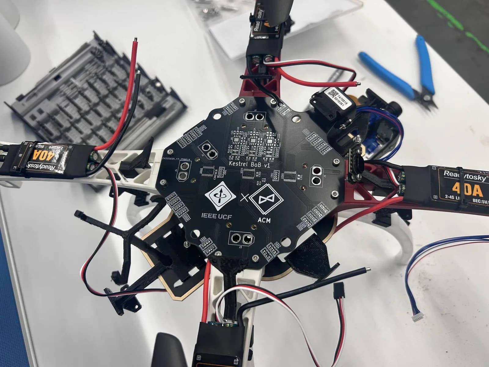 Top-down view of the black Kestrel BoB v1 circuit board, co-branded IEEE UCF and ACM, bolted to a red-and-white quadcopter frame among motor wiring and electronic speed controllers.