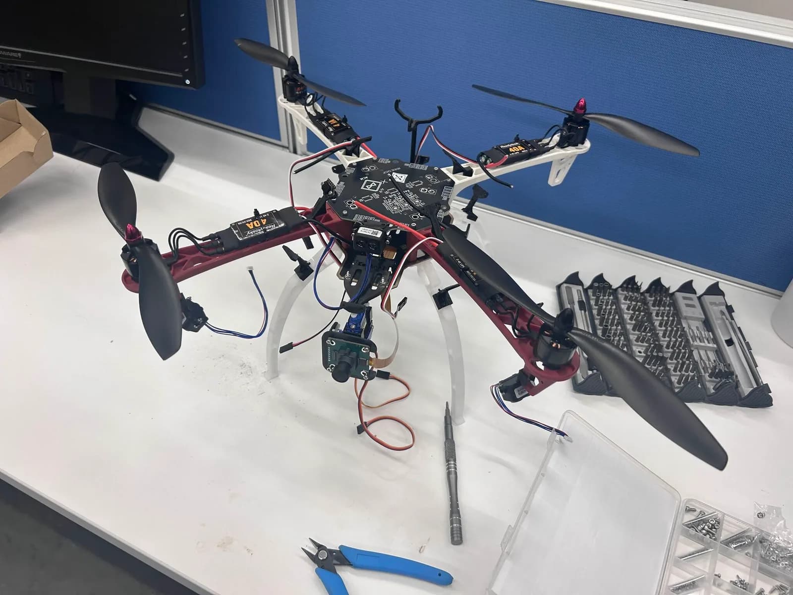 The assembled Kestrel quadcopter on a white workbench, with red arms, four black propellers, printed landing legs, and a downward-facing camera module.