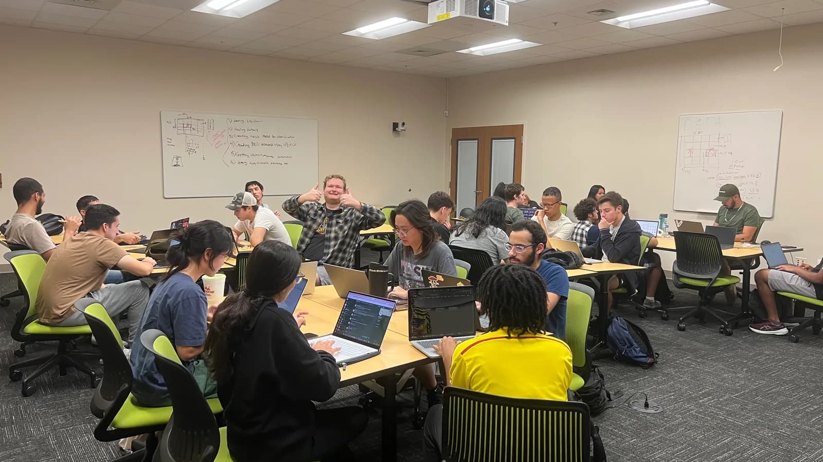 Kestrel members working on laptops around classroom tables, with a numbered agenda and system sketches on the whiteboards behind them.