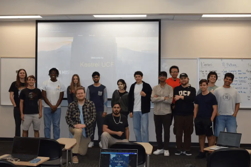 About twenty Project Kestrel members standing together in a classroom in front of a projected slide reading 'Welcome to Kestrel UCF'.