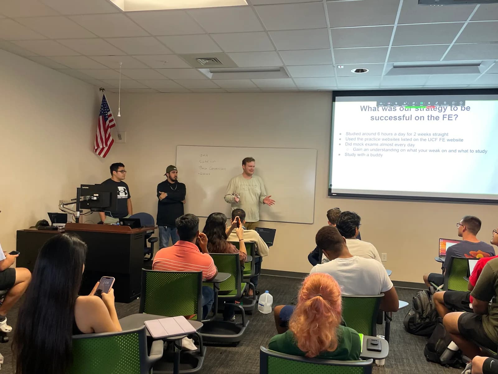 Three presenters at the front of a classroom in front of a projected slide titled 'What was our strategy to be successful on the FE?', speaking to a seated audience of students.